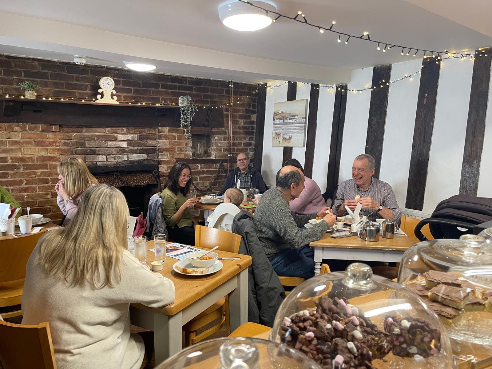 A busy afternoon in The Angel Café, with people eating and chatting at wooden tables beside the brick inglenook fireplace, under fairy lights and exposed timber beams